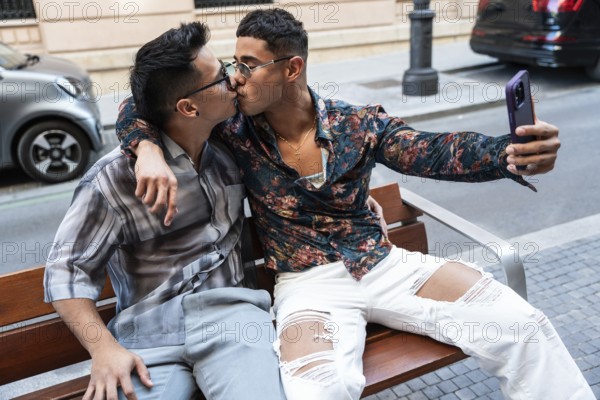 A latin gay couple shares a kiss while taking a selfie on a bench in Madrid. They display affection and joy in the bustling, urban setting, celebrating love openly