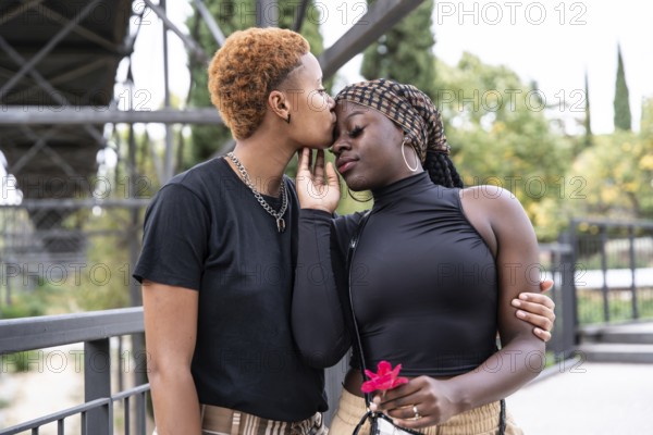 A loving LGBTQ multiethnic couple embraces in a park, sharing a tender moment. One partner gently kisses the other's forehead, exemplifying a genuine connection and affection