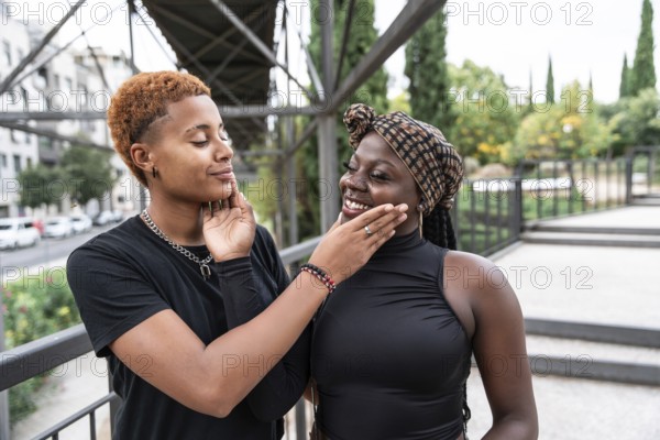A joyful LGBT multiethnic couple shares a tender moment outdoors, gently touching each other's faces. Surrounded by an urban setting, their expressions radiate love and happiness