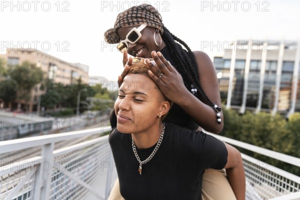 A joyful LGBT multiethnic couple shares playful moments outside. They enjoy each other's presence on a modern city bridge, surrounded by urban scenery and greenery