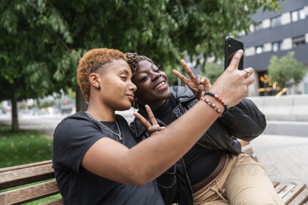 A joyful LGBT multiethnic couple taking a selfie on a park bench. They smile and flash peace signs, embodying happiness and connection. Lush greenery and urban scenery surround them