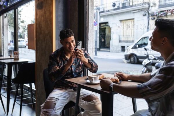 A latin gay couple enjoys coffee and pastries while sitting at a cafe. One partner is engaging with a smartphone, sharing a relaxed moment in a cozy urban setting