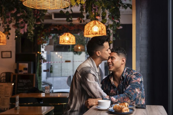 A latin gay couple shares a tender kiss in a warmly lit cafe surrounded by lush decor. They enjoy coffee and pastries, illustrating love and intimacy in a casual setting