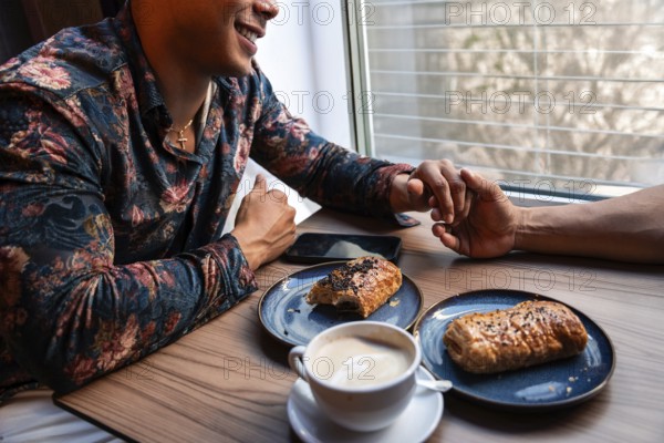 A latin gay couple shares a tender moment holding hands at a cafe. On the table are plates with pastries and a cup of coffee, creating a cozy and intimate ambiance