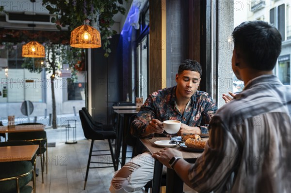 A latin gay couple enjoys a cozy moment at a café, sharing coffee and pastries. The warm lighting and modern decor create a relaxed and intimate atmosphere