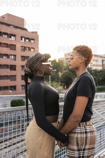 A loving LGBT multiethnic couple stands closely, facing each other, on an outdoor walkway. The setting sun casts a warm glow, highlighting the modern urban backdrop and their connection