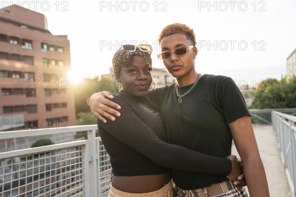 A loving LGBT multiethnic couple embraces outdoors on a bridge during a beautiful sunset. The warm light enhances their affectionate pose, symbolizing unity and love in an urban setting