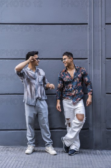 A joyful, stylish latin gay couple shares a lively conversation against a modern urban backdrop. Their casual fashion and genuine smiles capture a moment of connection and happiness