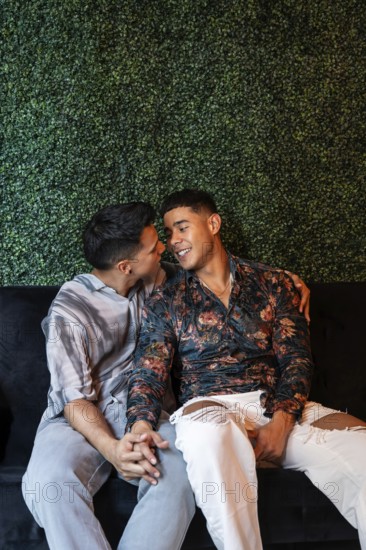 A loving latin gay couple sits closely on a sofa, holding hands and sharing a joyful, intimate moment with smiles, against a lush green plant wall, expressing warmth and affection