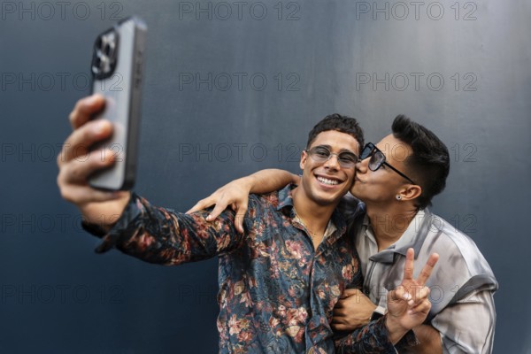 A joyful latin gay couple takes a selfie, capturing a moment of affection and happiness. One person kisses the other on the cheek, while making a peace sign