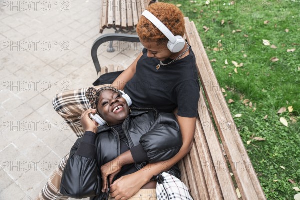 A joyful LGBT multiethnic couple relaxes on a park bench, listening to music through headphones. Their smiles and closeness depict love and happy companionship on a sunny day
