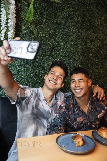 A joyful latin gay couple takes a selfie while enjoying brunch at a café. Their smiles and stylish outfits reflect a vibrant, loving atmosphere, surrounded by lush green decor