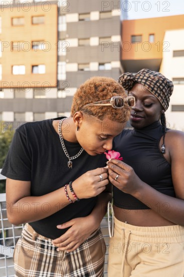 A joyful LGBT multiethnic couple shares a tender moment outdoors. One person gently sniffs a flower, while the other stands close with a warm smile. Urban background and sunny weather
