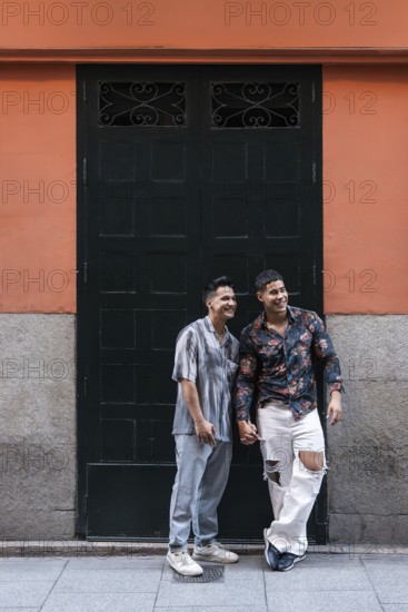 A joyful latin gay couple holds hands while standing in front of a colorful building in Madrid. The scene captures love, diversity, and cultural vibrancy