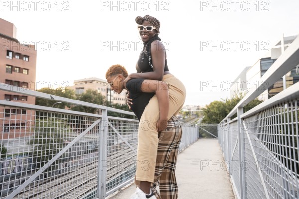 A happy LGBT multiethnic couple enjoys a playful moment on a bridge in an urban setting, with one partner giving the other a piggyback ride. The sky is clear and the mood is cheerful