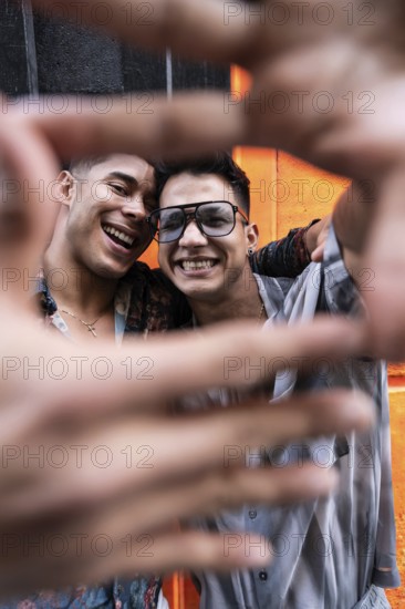 A joyful latin gay couple poses with smiles, framing their faces with their hands for a selfie against a vibrant orange wall, capturing a moment of happiness and love