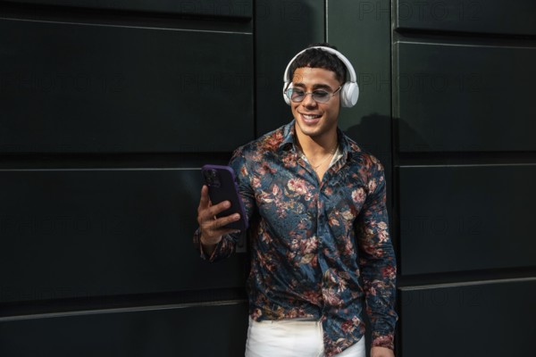 A fashionable man in sunglasses and a floral shirt stands against a dark green wall, wearing headphones and holding a smartphone, enjoying music outdoors