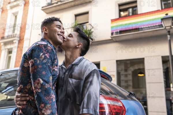 A joyful latin gay couple shares a tender moment on a lively street in Madrid, with a rainbow flag in the background, symbolizing pride and love