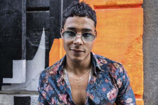 A confident latin man wearing sunglasses and a floral shirt poses against a vibrant, colorful graffiti wall, showcasing a bold, urban style with a casual yet trendy look