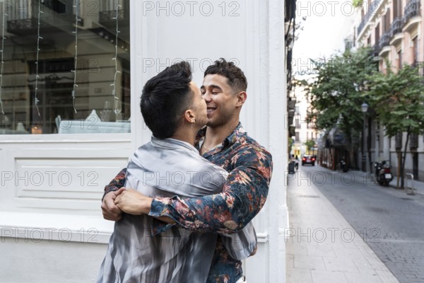 A joyful latin gay couple embraces and shares a tender moment on a charming street in Madrid. The vibrant city backdrop adds to the warmth of their connection and happiness