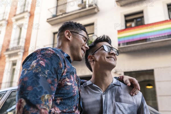 A joyful latin gay couple embraces, smiling on the vibrant streets of Madrid. The background features a rainbow flag, adding to the celebratory atmosphere