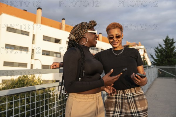 A happy LGBT multiethnic couple smiling while holding phones, enjoying the sunny outdoors in fashionable casual clothing, standing on a bridge with modern buildings in the background