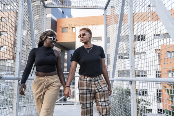 A fashionable LGBT multiethnic couple walks hand in hand across a modern footbridge, smiling and enjoying their time together. Urban architecture frames their joyful interaction