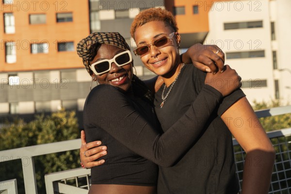 A happy LGBT multiethnic couple embraces warmly outdoors, both smiling widely. They wear stylish sunglasses and casual attire, with urban buildings in the background