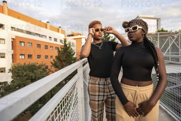 A fashionable LGBT multiethnic couple poses confidently on an urban rooftop, showcasing their unique style with sunglasses. The modern cityscape adds a vibrant background