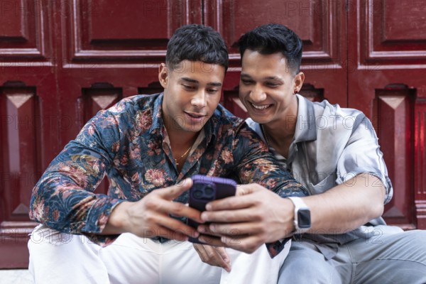 A joyful latin gay couple shares a happy moment in Madrid city. They are sitting together, smiling and looking at a phone screen, reflecting connection and love