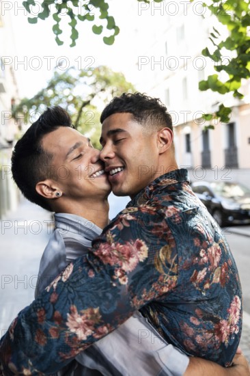 A joyful latin gay couple embraces lovingly on a sunny street in Madrid. The background features charming city architecture and lush greenery, highlighting the vibrant urban setting