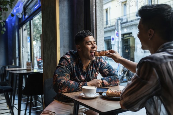 A joyful latin gay couple shares a pastry at a cafe, highlighting a moment of connection. The atmosphere is cozy and inviting, portraying warmth and happiness