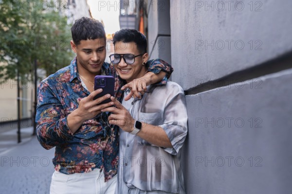 A joyful latin gay couple stands close together on a sunny street in Madrid, sharing a moment as they look at a smartphone screen. The scene captures happiness and companionship