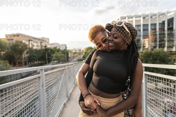 A happy LGBT multiethnic couple embraces and smiles warmly at each other on an urban bridge, with a cityscape backdrop during a sunny day, expressing love and joy