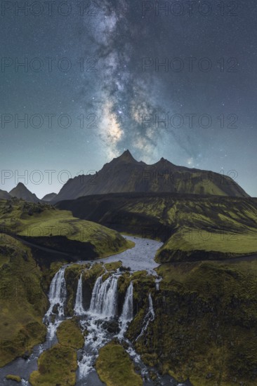 A breathtaking view of the Milky Way illuminating the night sky over a lush Icelandic landscape, featuring a cascading waterfall and rugged mountains under the starry expanse