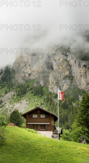 A cozy wooden chalet stands amidst lush greenery, with misty cliffs in the background at Lac de Taney, Switzerland A Swiss flag waves gently in the breeze