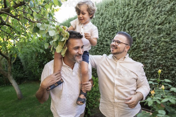 A joyful moment captured outdoors shows a gay couple spending quality time with their young son. The family is surrounded by lush greenery, embodying love and togetherness