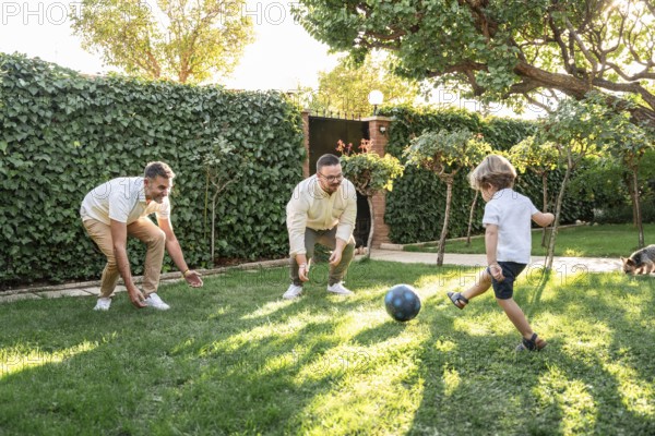 A happy gay couple plays soccer with their young son in a sunlit garden. The lush greenery and relaxed atmosphere highlight love, family, and shared moments outdoors