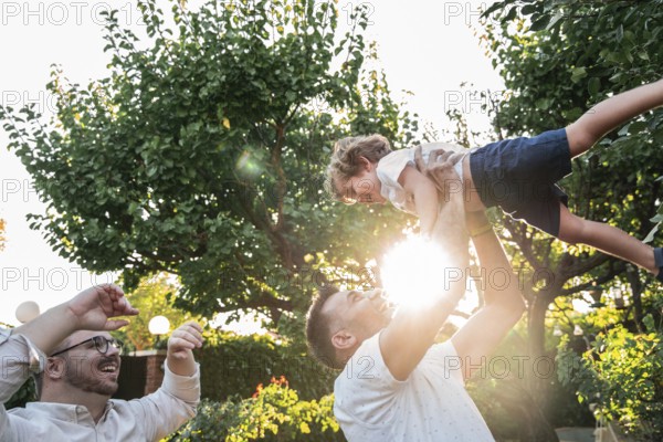 A happy gay couple plays with their young son outdoors on a sunny day, surrounded by lush greenery. The child is lifted in the air, enjoying quality family time