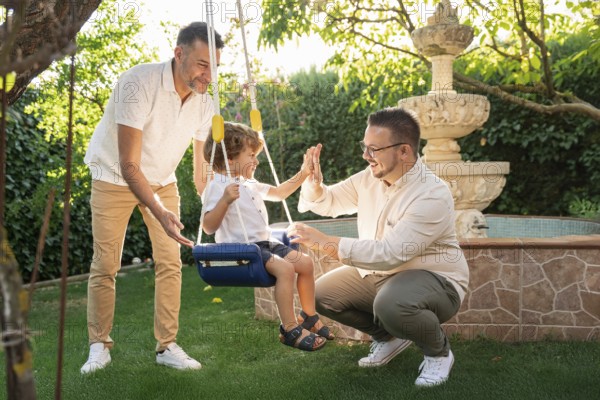 A joyful gay couple engages with their young son in a sunny garden, sharing laughter and high-fives while he swings, embodying love, happiness, and family togetherness