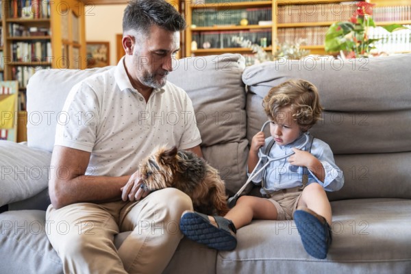A boy with a stethoscope playfully examines a dog while sitting on a couch. His grandfather watches with a smile in their cozy living room filled with books and plants