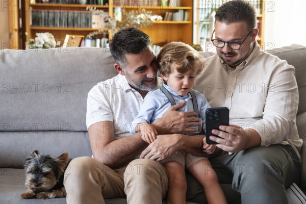 A joyful gay couple sits on a cozy couch with their young son and a small dog, sharing moments together while looking at a smartphone, symbolizing modern family love