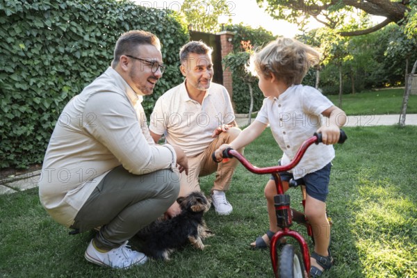 A joyful gay couple engages with their young son in a vibrant green garden, surrounded by lush foliage. One child rides a red bicycle, capturing a moment of family bliss