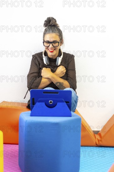 A woman teaching sign language through a video call, sitting on colorful cushions with a tablet Headphones rest around her neck, creating a lively learning atmosphere