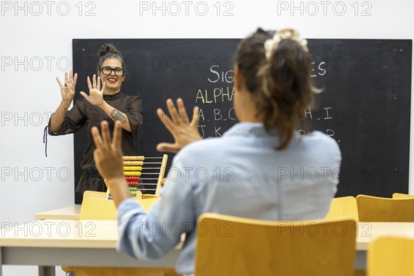 A sign language instructor teaches a student in a classroom The student mirrors hand gestures while seated at a desk, and a blackboard displays the sign alphabet