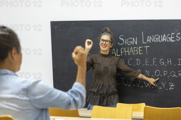 An instructor teaches sign language, pointing to an alphabet chart on a chalkboard A student practices hand signs, fostering communication skills and inclusivity