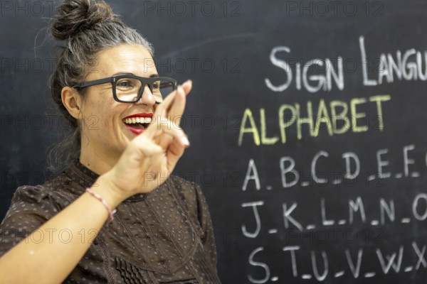 A woman teaches sign language alphabet in a classroom, gesturing with a smile Chalkboard filled with letters in the background adds an educational atmosphere