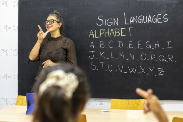 A sign language instructor demonstrates letters from the alphabet on a blackboard in a classroom setting Students engage by practicing signs in a bright, welcoming environment