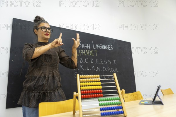 An instructor demonstrates sign language, using hand gestures to teach the alphabet The classroom setting includes a chalkboard with letters and an abacus on the table