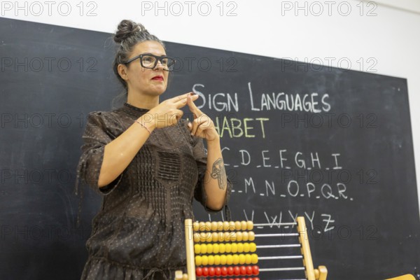 An instructor teaching sign language in a classroom, standing in front of a chalkboard with the alphabet written on it An abacus is visible in the foreground